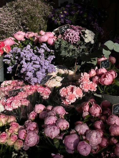 A vibrant floral display featuring an assortment of fresh flowers arranged in bouquets and clusters, including soft pink peonies, purple statice, white blooms, and sprigs of eucalyptus. Some flowers a