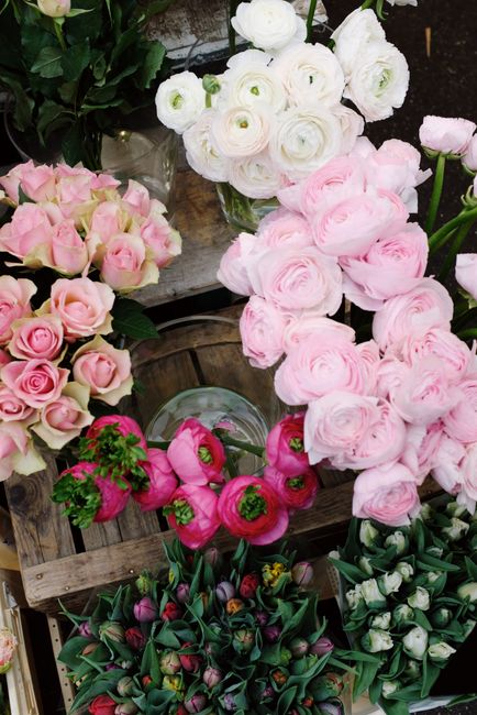 An assortment of fresh flowers displayed at a flower stall, featuring a variety of pastel pink roses, white ranunculus with delicate, layered petals, and vibrant pink tulips. The flowers are arranged 