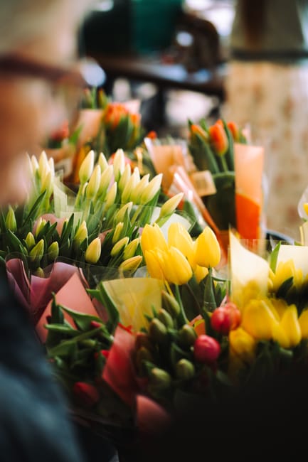 A close-up of a vibrant selection of fresh flowers arranged in bouquets at a florist shop, featuring yellow tulips, white tulips, and pink blossoms wrapped in pastel-colored paper. The flowers are dis