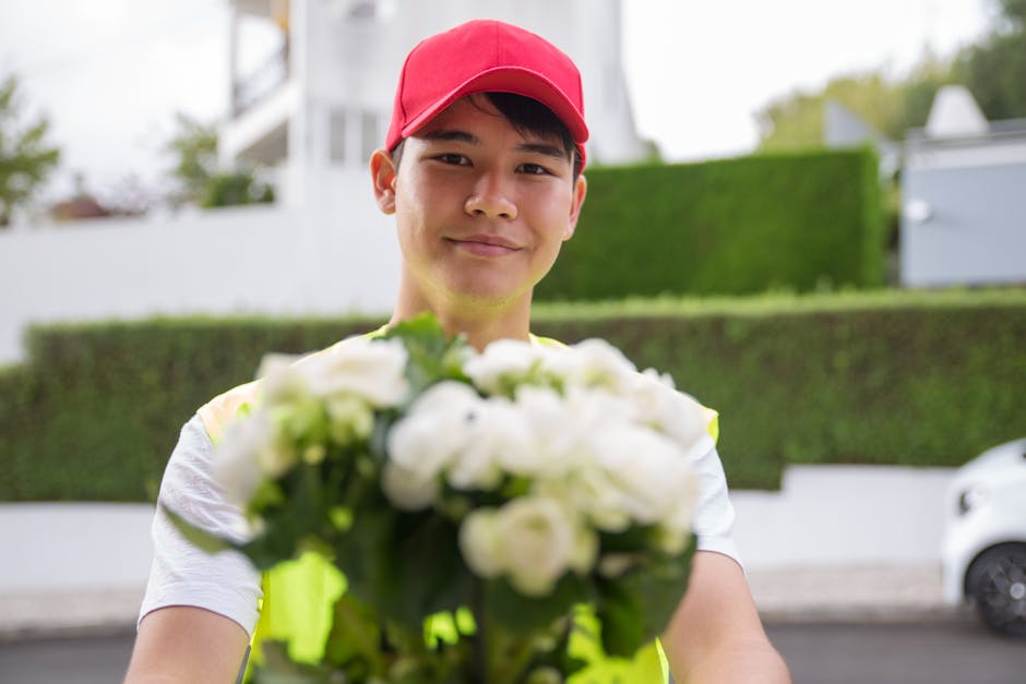 A smiling delivery person in a red cap and light-colored t-shirt holding a bouquet of white flowers with green leaves, extending the floral arrangement toward the camera. The bouquet is fresh, with de