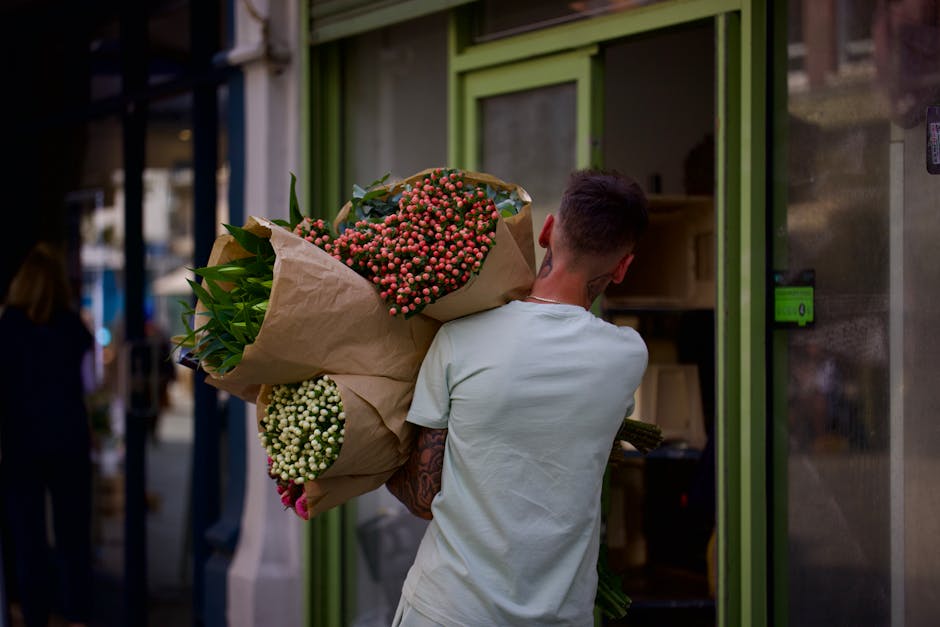 A person with short dark hair, wearing a light-colored T-shirt, is holding a large bouquet of fresh flowers wrapped in brown kraft paper. The bouquet contains pink and white berries, green foliage, an