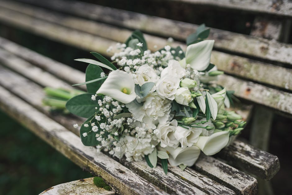 A beautifully arranged wedding bouquet featuring white roses, calla lilies, and small baby's breath flowers, accented with lush green foliage, resting on a weathered wooden bench outdoors. The bouquet