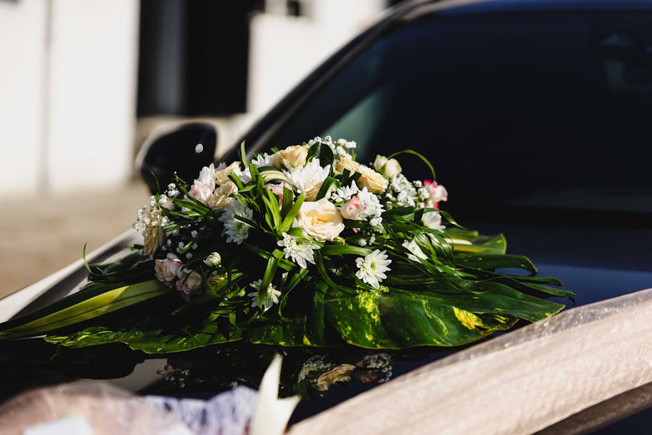 A delicate floral arrangement placed on the hood of a black vehicle, featuring a bouquet of fresh white roses, small white daisies, and baby's breath, all accented with lush green foliage and long lea