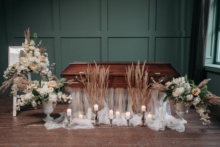 A funeral floral display arranged on a wooden floor in front of a closed wooden coffin against a dark green paneled wall. The arrangement includes two large white vases filled with cream and white ros